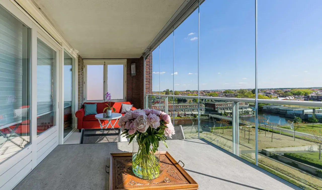 Een prachtig uitzicht vanaf dit balkon, dat met glazen schuifwanden is dichtgemaakt. Een rode bank en bloemen maken het gezellig.