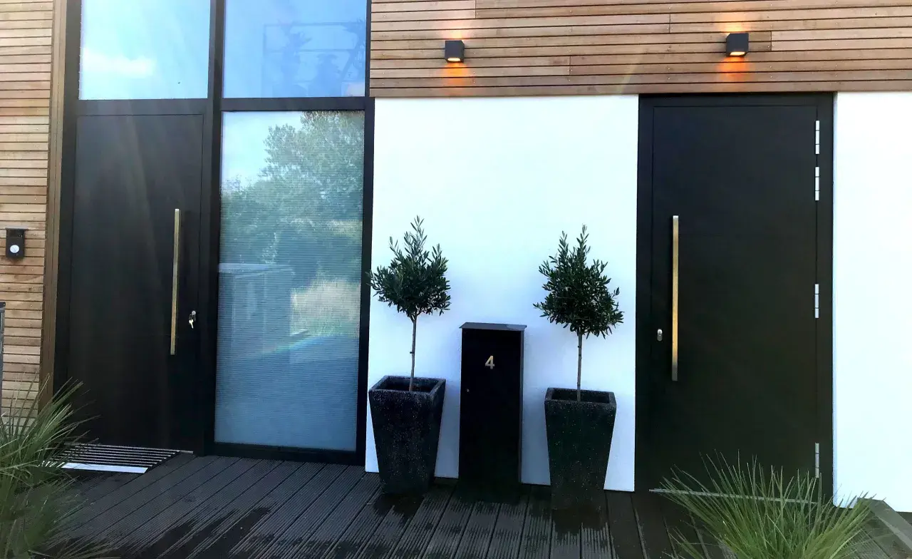 Moderne zwarte voordeur met gouden handgreep, naast een groot raam. Twee olijfbomen in potten en een zwarte brievenbus met nummer 4 bieden voordeur inspiratie.
