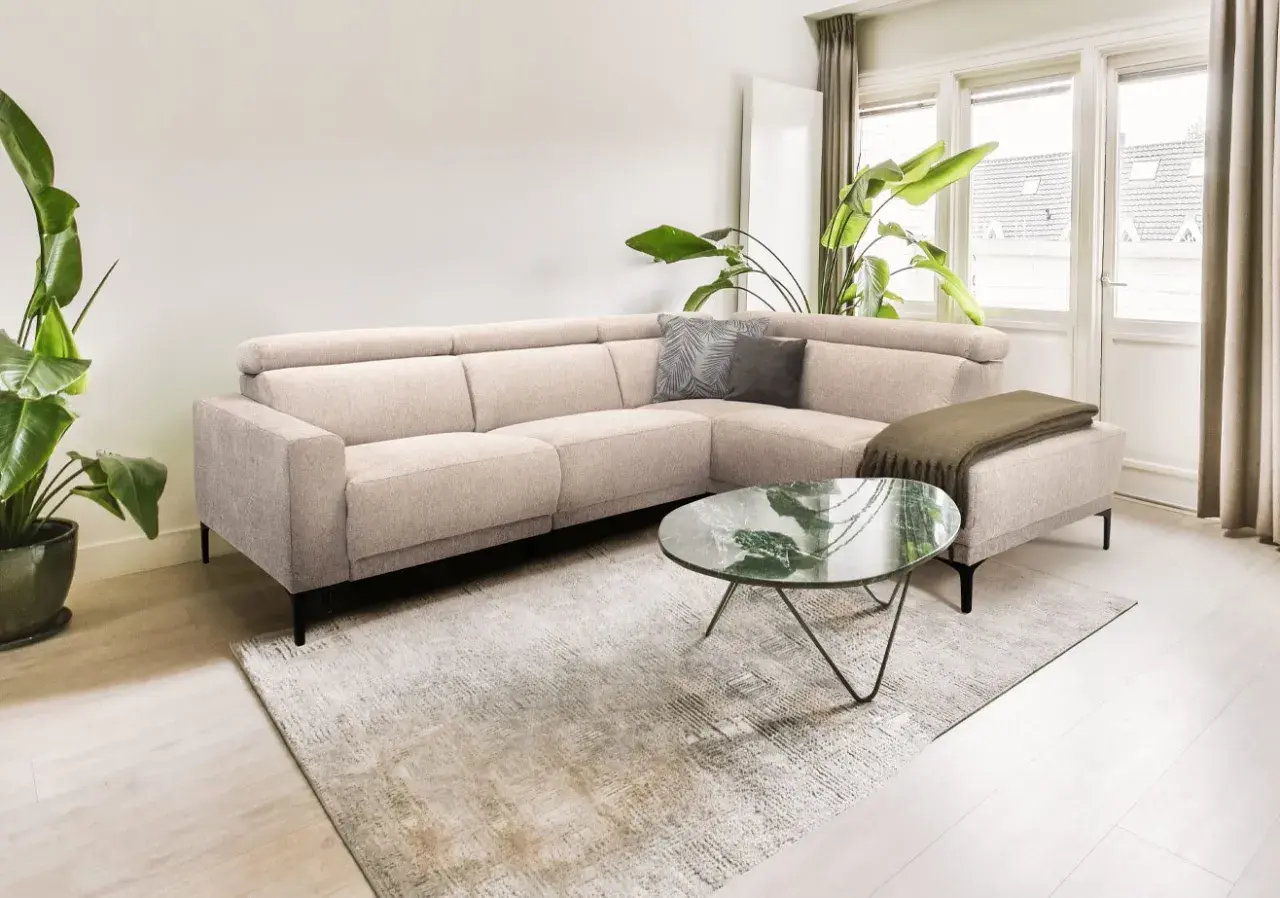 Licht beige hoekbank met kussens en plaid, een marmeren salontafel en grote planten creëren een rustig slow living interieur.