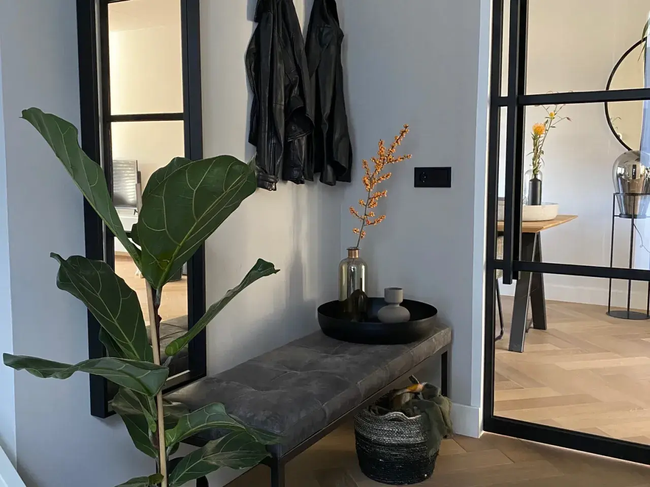 Een mooie hal met een grote plant, leren jasjes aan de kapstok, een bankje en een mand.