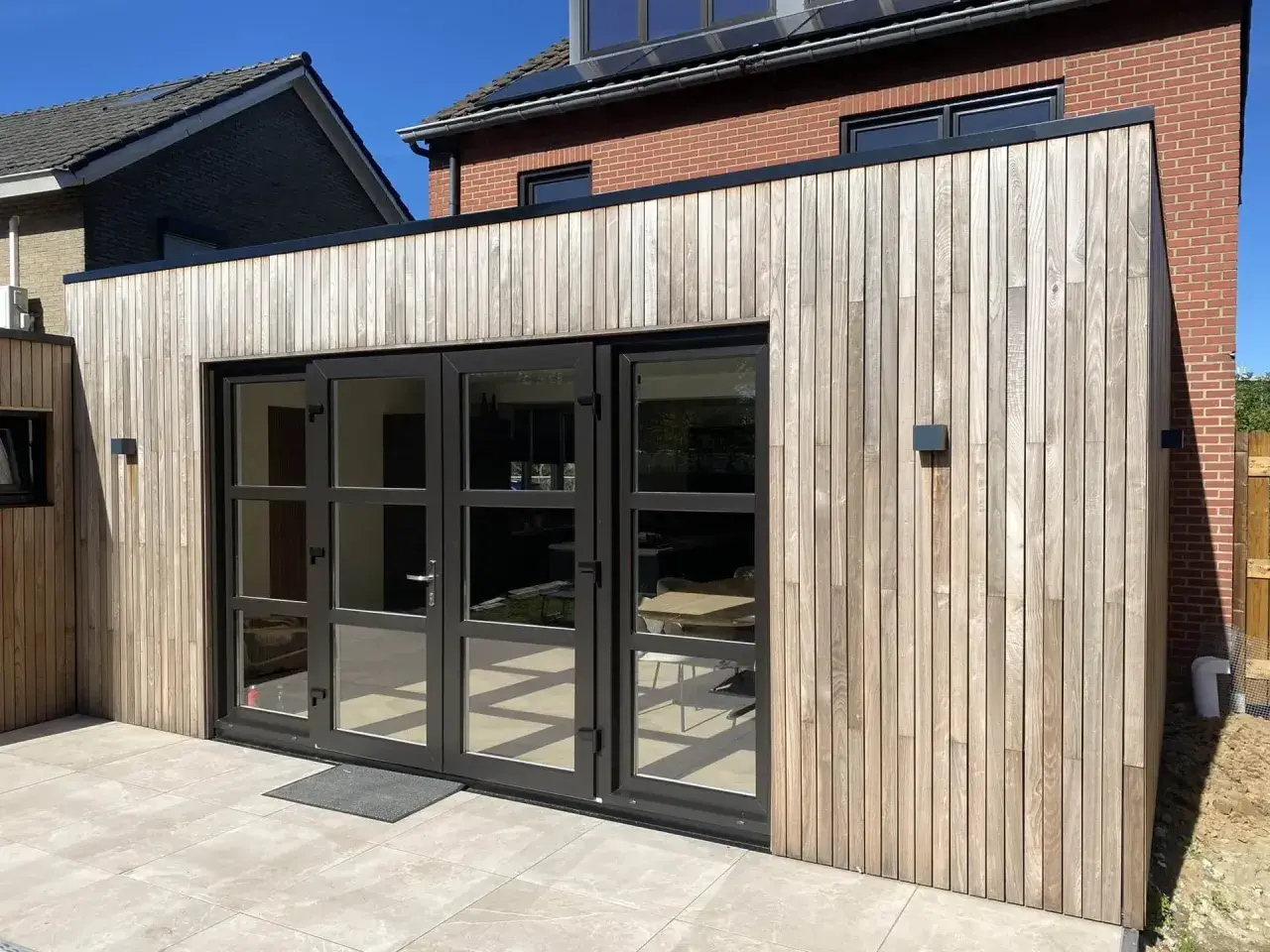 Moderne uitbouw voorkant huis met houten gevelbekleding en grote glazen deuren. De zon werpt schaduwen op de lichte terrastegels.