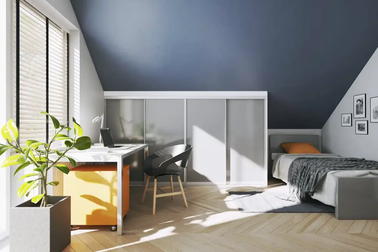 Een smalle hal met een bureau, bed en kasten. De zon schijnt door het raam en werpt patronen op de houten vloer.
