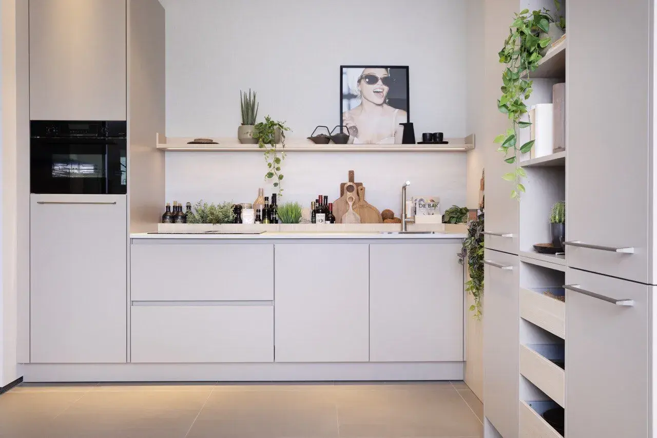 Moderne nis keuken met ingebouwde oven, strakke kasten en decoratieve planten. Een poster met een lachende vrouw siert de muur.