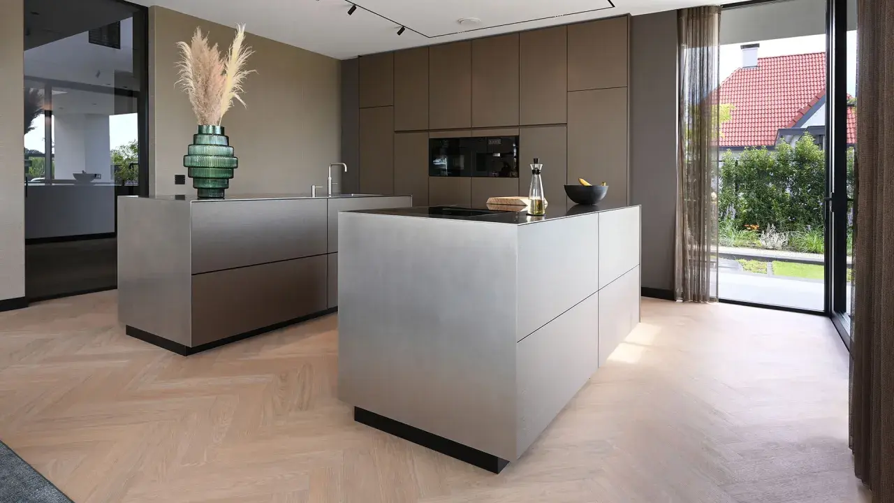 Moderne keuken met strakke, metalen kasten op een visgraat houten vloer. Een groene vaas met pampasgras siert het aanrecht.