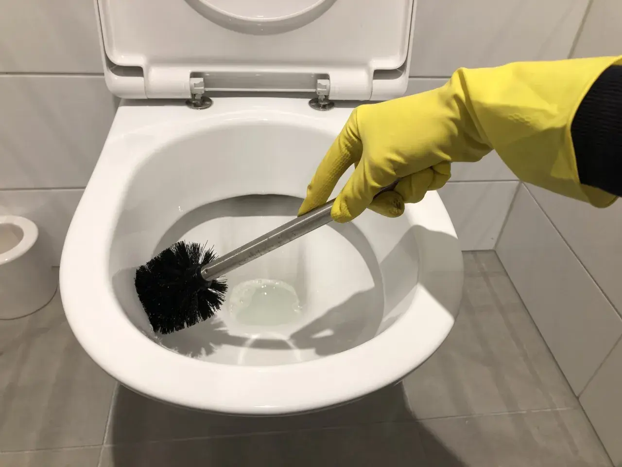 Hand met gele handschoen is bezig met wc poetsen. De toiletborstel maakt de pot schoon.