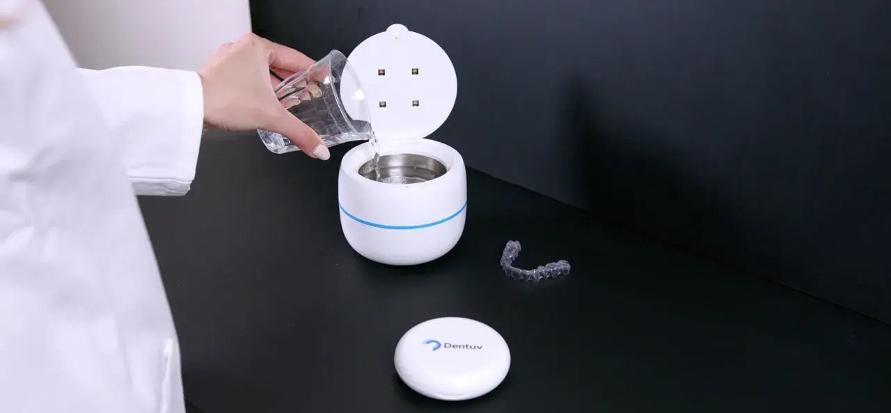 Een hand met een glas water vult een apparaat voor ultrasoon reinigen. Een bitje ligt ernaast.