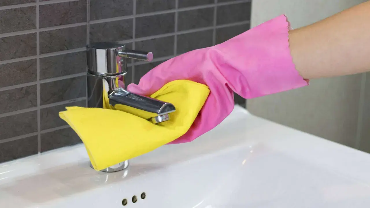 Hand in roze handschoen maakt de kraan van de wasbak schoon met een gele doek.
