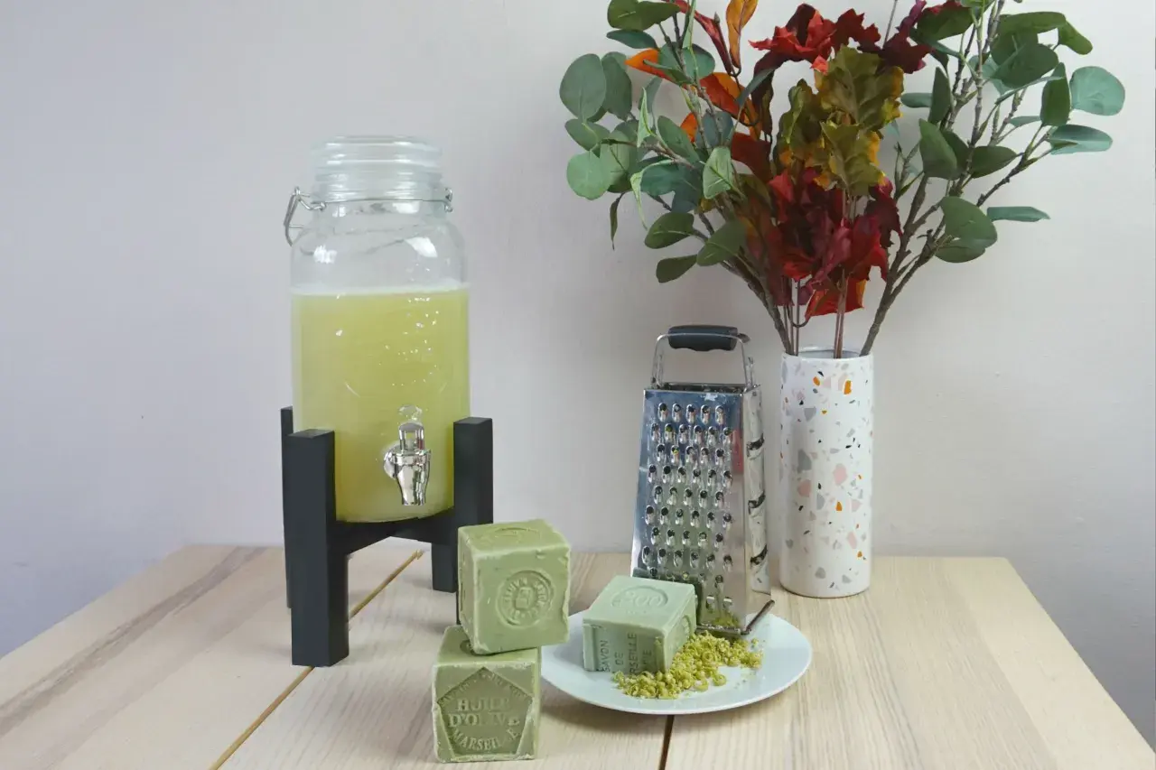 Zelf wasmiddel maken ervaringen: groene zeepblokken, rasp, en een glazen dispenser met gele vloeistof. Een vaas met herfstbladeren completeert het tafereel.