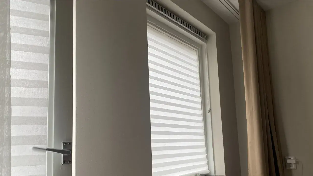 Wit plisségordijn als tijdelijke raambekleding tijdens een verbouwing. Een beige gordijn hangt ernaast.
