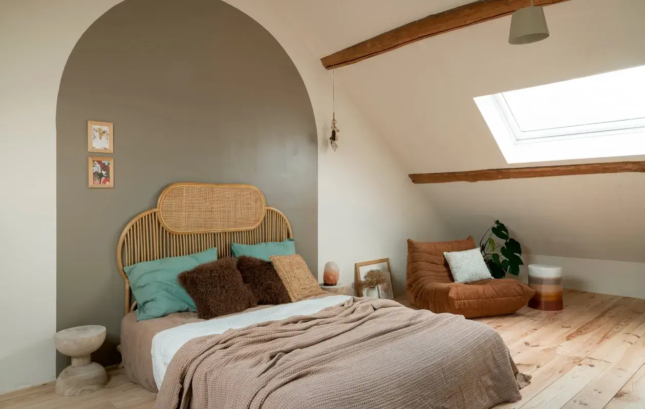 Een rustige, minimalistische slaapkamer met een rieten hoofdeinde, zachte plaids en een comfortabele fauteuil onder een dakraam.