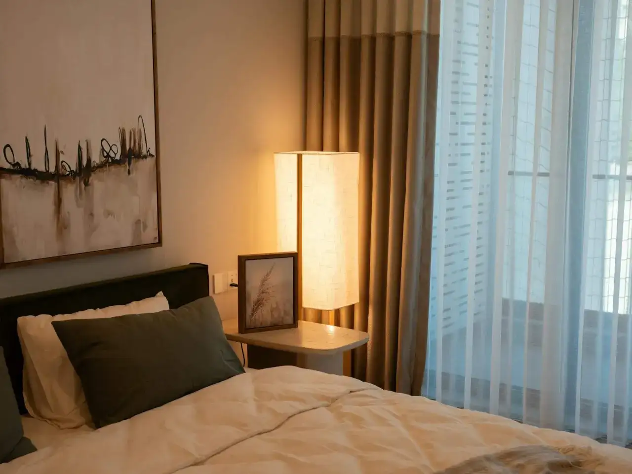 Een comfortabel bed voor een raam met lichte gordijnen. Een staande lamp en een schilderij aan de muur creëren een rustige sfeer.