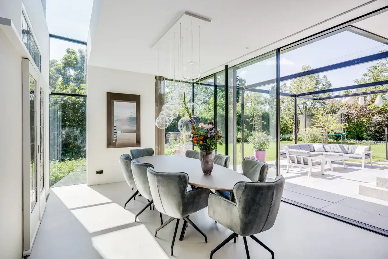 Een lichte, moderne eetkamer met grote ramen die uitkijken op de tuin. De eettafel is omringd door comfortabele stoelen, wat een uitnodigende vestibule cre&euml;ert.