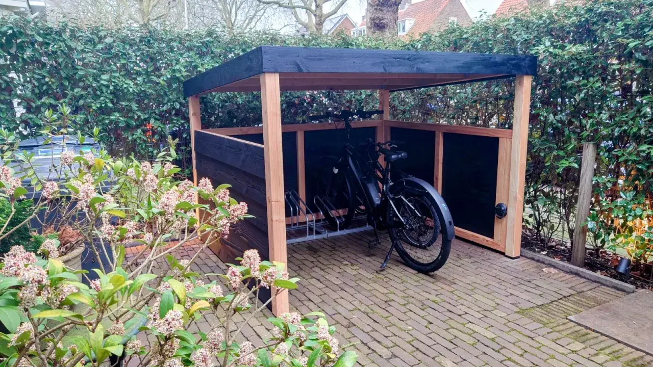 Een nette oplossing voor fietsen in de voortuin: een houten fietsenstalling met zwarte wanden en dak, waarin twee fietsen staan.