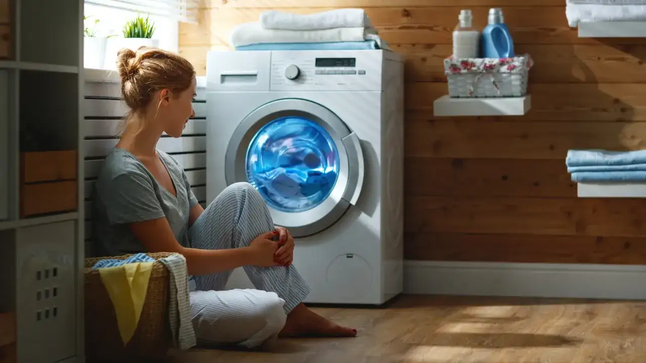 Vrouw kijkt naar draaiende wasmachine met handdoeken en ondergoed. Een mand met wasgoed staat naast haar.
