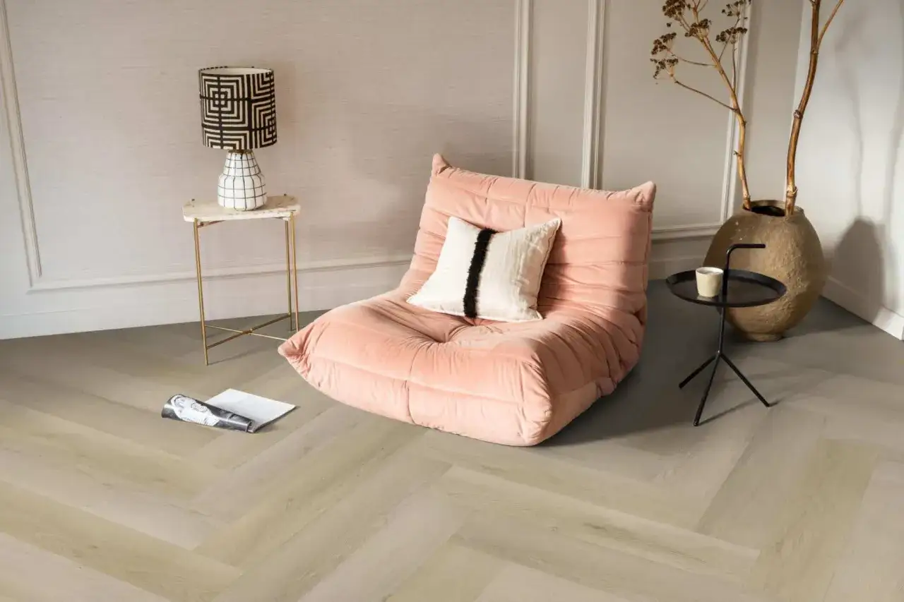 Een roze fauteuil staat op een lichte evc vloer met visgraatmotief. Een bijzettafeltje met lamp en een tijdschrift completeren de sfeer.