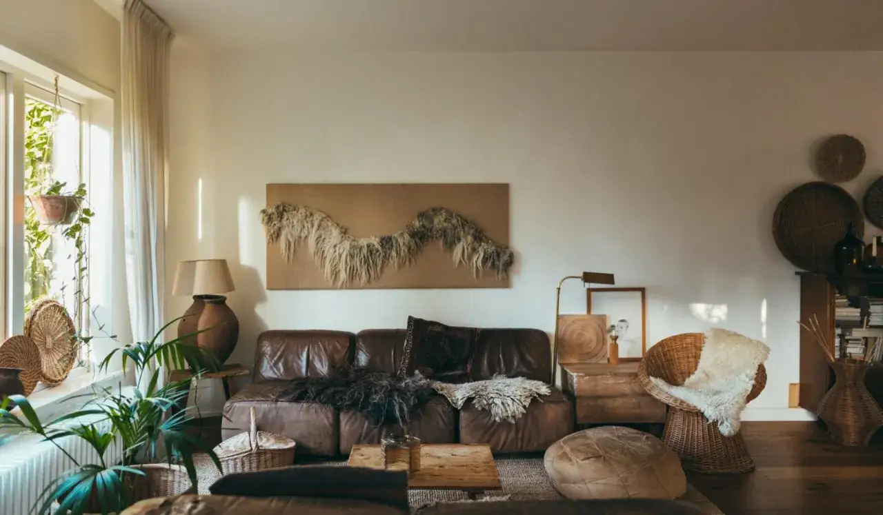 Gezellige woonkamer met bruine lederen bank, rieten stoel en planten. Een kunstwerk aan de muur en manden dragen bij aan de sfeer. Perfect voor goedkoop verbouwen met een bohemian touch.