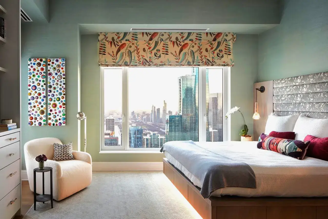 Een modern bed voor raam met uitzicht op de skyline. Een comfortabele fauteuil en kleurrijke kunst aan de muur maken de kamer compleet.