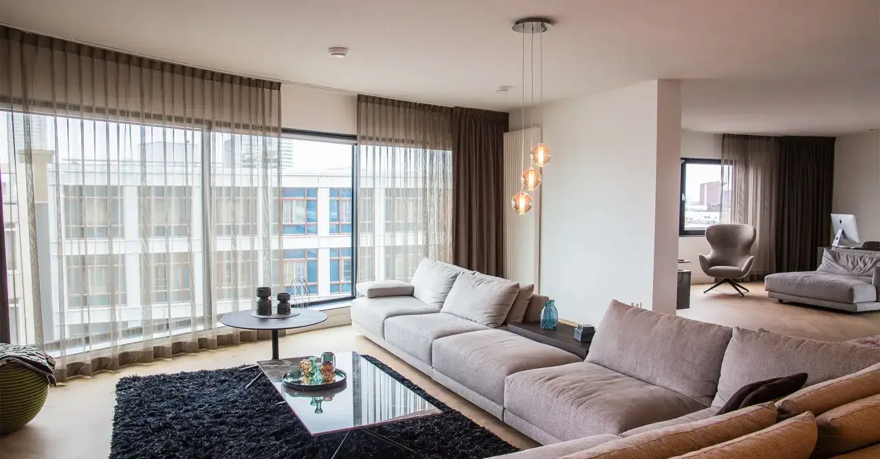 Luxe woonkamer met grote hoekbank, glazen salontafel en een nis in de muur. Grote ramen met uitzicht op de stad.