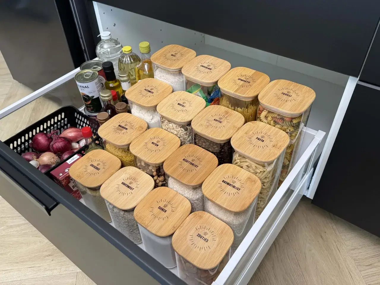 Netjes opgeborgen plastic bakjes met bamboe deksels, gevuld met pasta, rijst, meel en muesli. Een georganiseerde keuken!