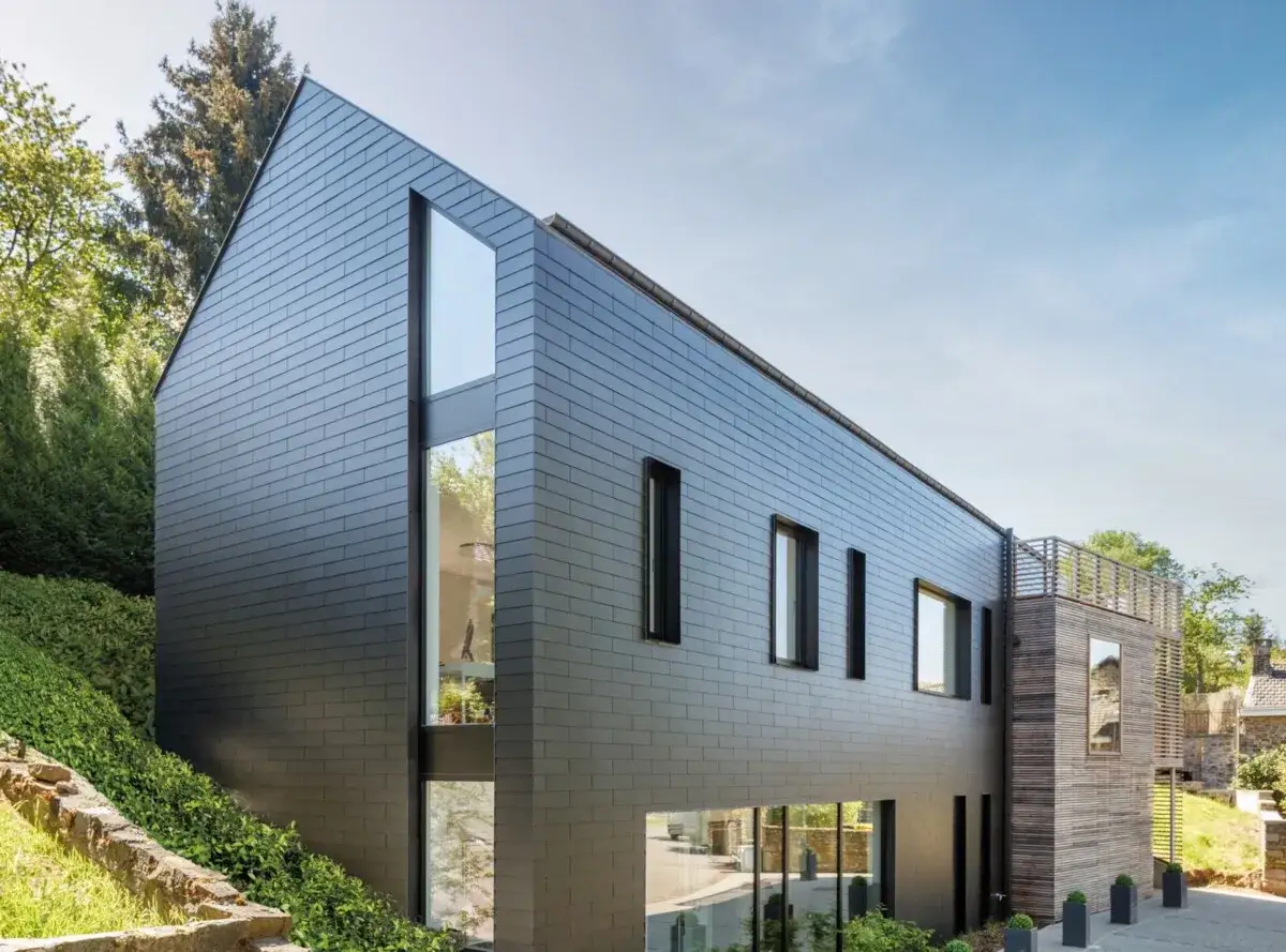 Moderne woning met een nieuwe gevel van donkere leien. Grote ramen bieden uitzicht op de groene omgeving.