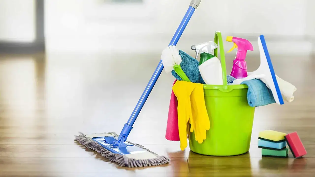 Een groene emmer met schoonmaakspullen, waaronder handschoenen, spuitflessen en een borstel, naast een dweil. Dit is een handig schoonmaakschema voor schoonmaakster.