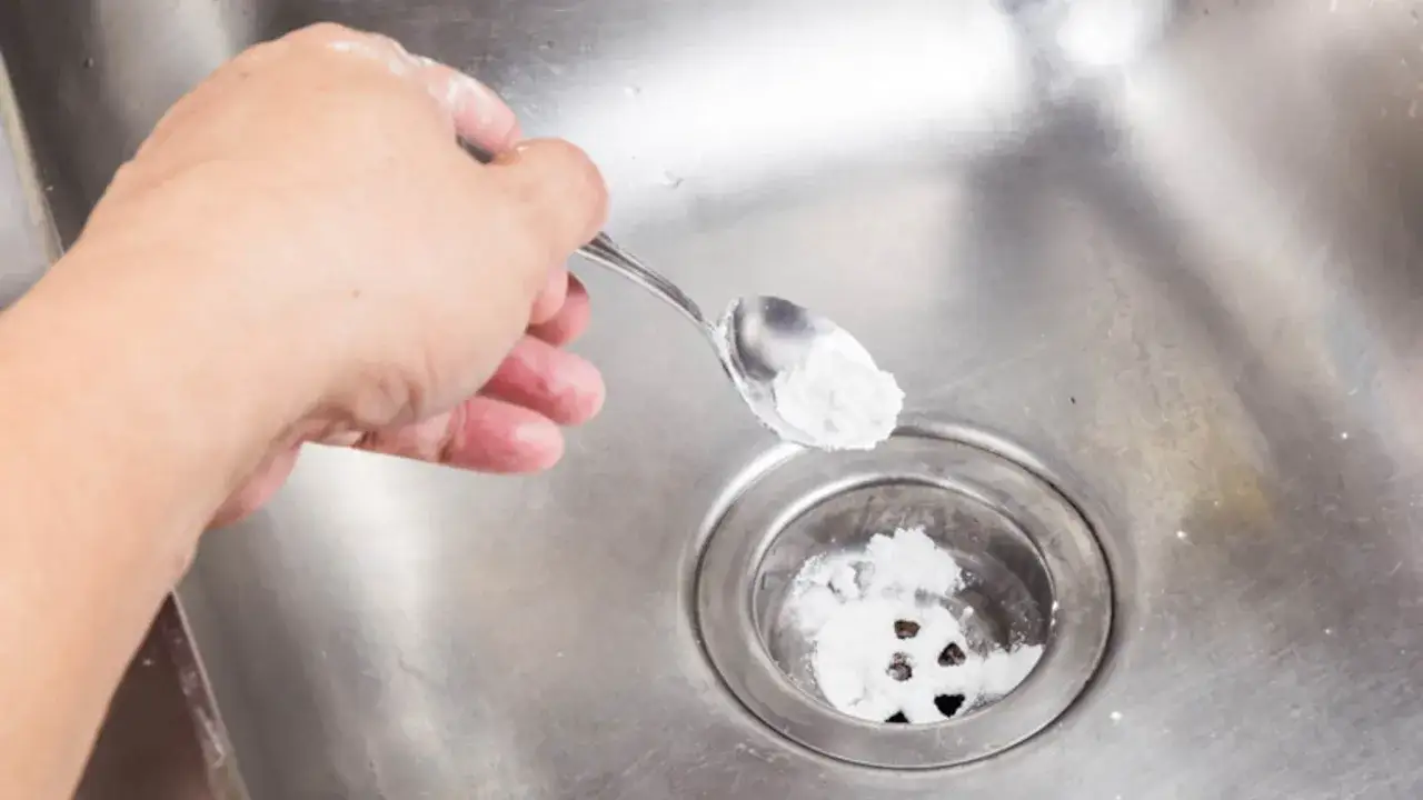 Een hand strooit met een lepel baking soda in een gootsteenafvoer. Dit is een stap in het wasbak schoonmaken.