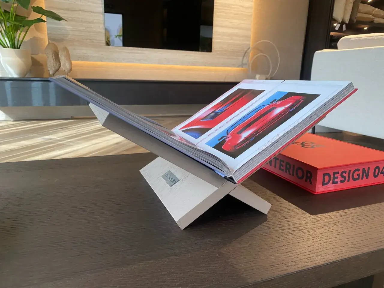 Een open boek met rode sportauto's rust op een stijlvolle, zelfgemaakte boekensteun. Naast een rode designboek.
