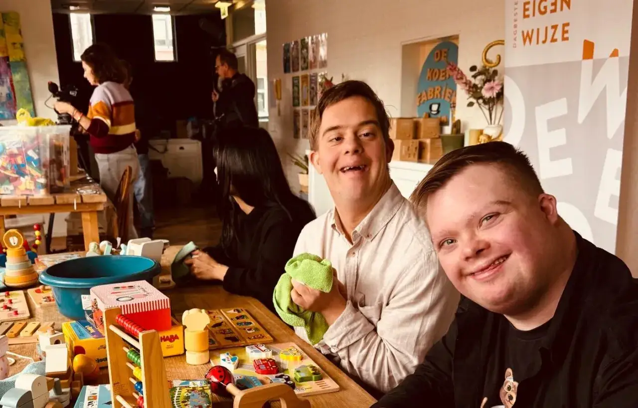 Twee lachende mannen met het syndroom van Down, omringd door speelgoed. Ze lijken te genieten van het moment, misschien wel na het opruimen of voorbereiden van speelgoed weggooien.