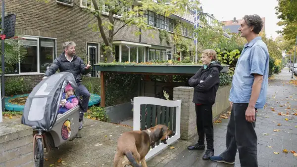 Een vader met dochter in bakfiets, zoon en hond staan buiten. Een slimme oplossing voor fietsen in de voortuin, met een groen dak boven de stalling.