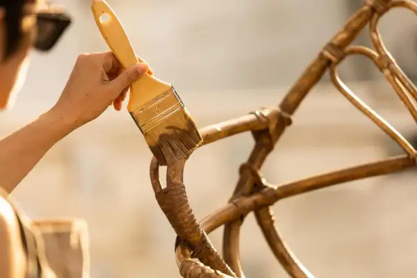 Hand met kwast werkt aan het **zelf rotan stoel repareren**. De stoel krijgt een opknapbeurt.