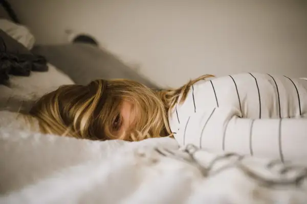 Een kind met blond haar ligt in bed, met een oog dat net zichtbaar is. De kamer is rommelig, het kind kan niet opruimen.