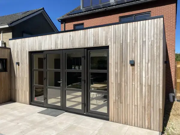 Moderne uitbouw voorkant huis met houten gevelbekleding en grote glazen deuren. De zon werpt schaduwen op de lichte terrastegels.