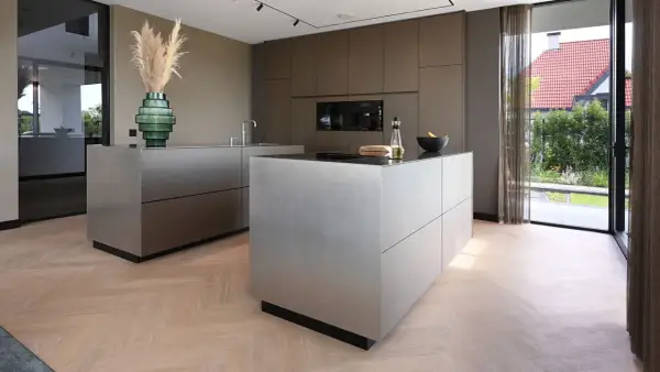 Moderne keuken met strakke, metalen kasten op een visgraat houten vloer. Een groene vaas met pampasgras siert het aanrecht.