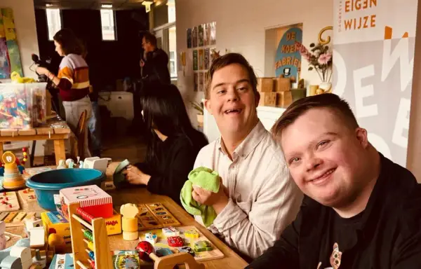 Twee lachende mannen met het syndroom van Down, omringd door speelgoed. Ze lijken klaar om het speelgoed op te ruimen, een gezellige activiteit.