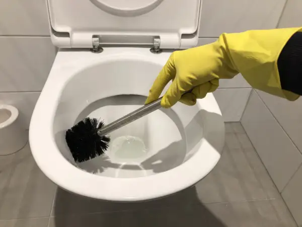 Hand met gele handschoen is bezig met wc poetsen. De toiletborstel maakt de pot schoon.
