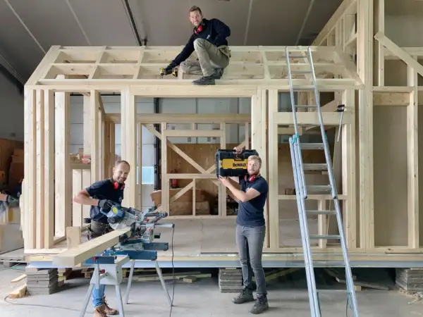 Drie mannen werken aan een **tiny huis bouwen**. Eén zaagt hout, een ander schroeft aan het dak, en de derde draagt een gereedschapskist.